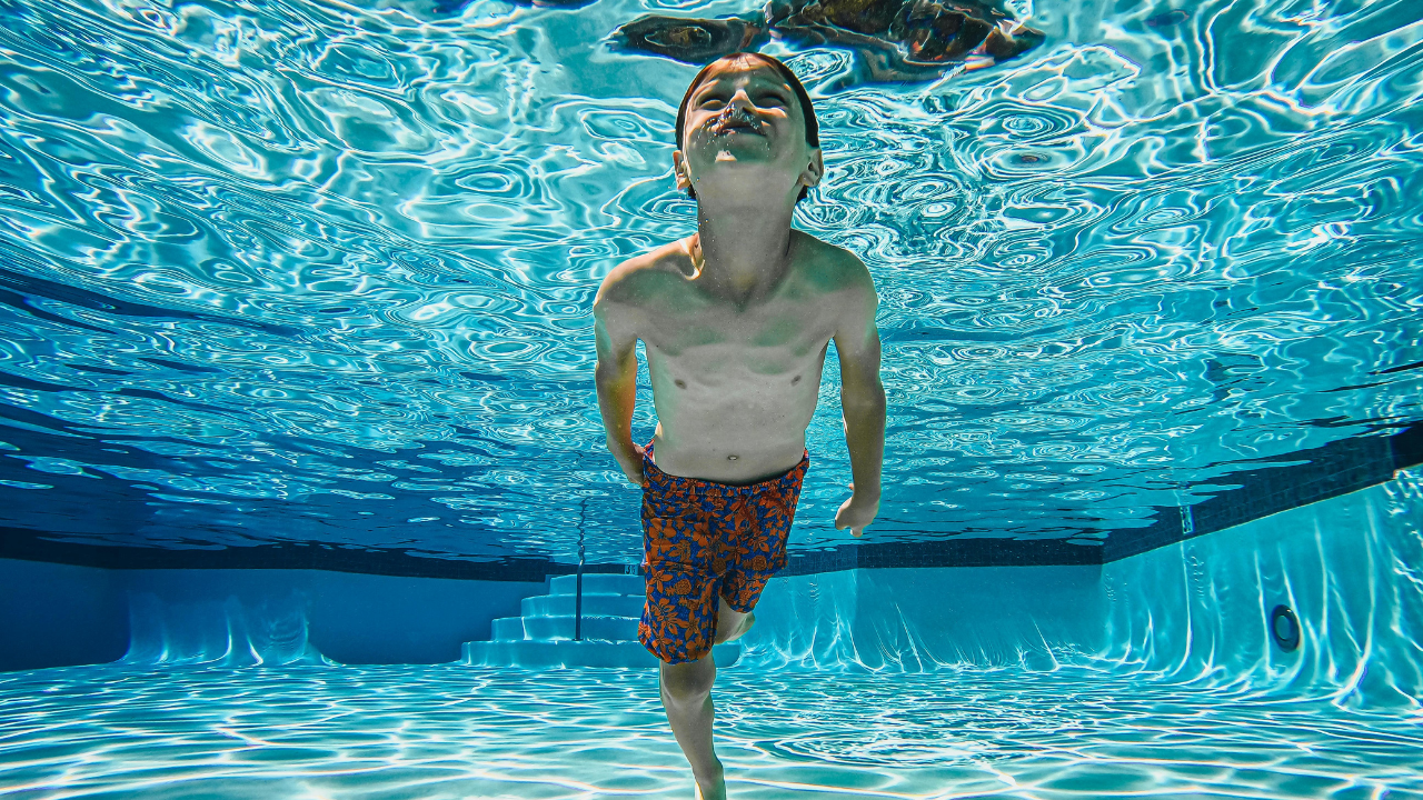 A little boy in blue floral swimming shorts submerged underwater in a pool, looking up.