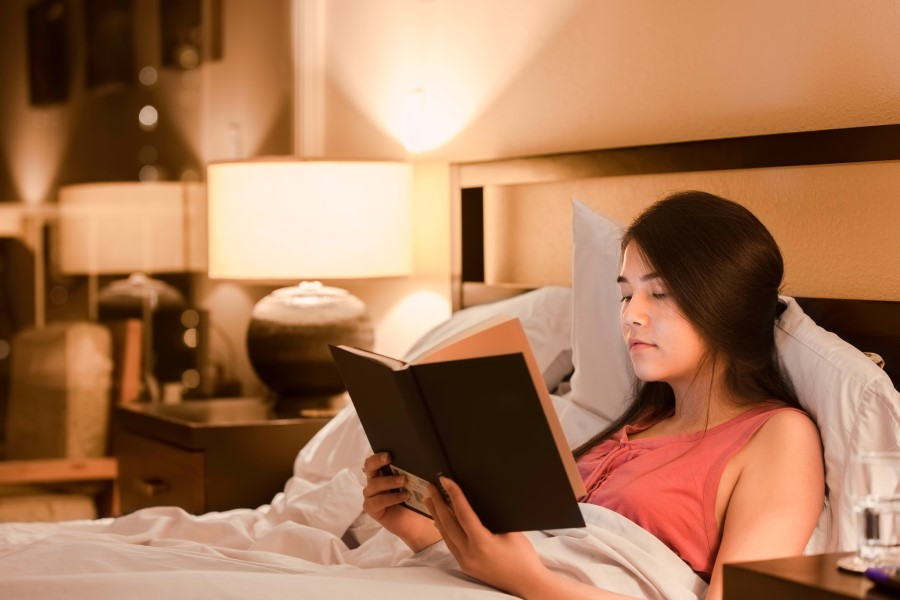 A woman is reading a book before bedtime surrounded by the soft glow of her bedside lamp